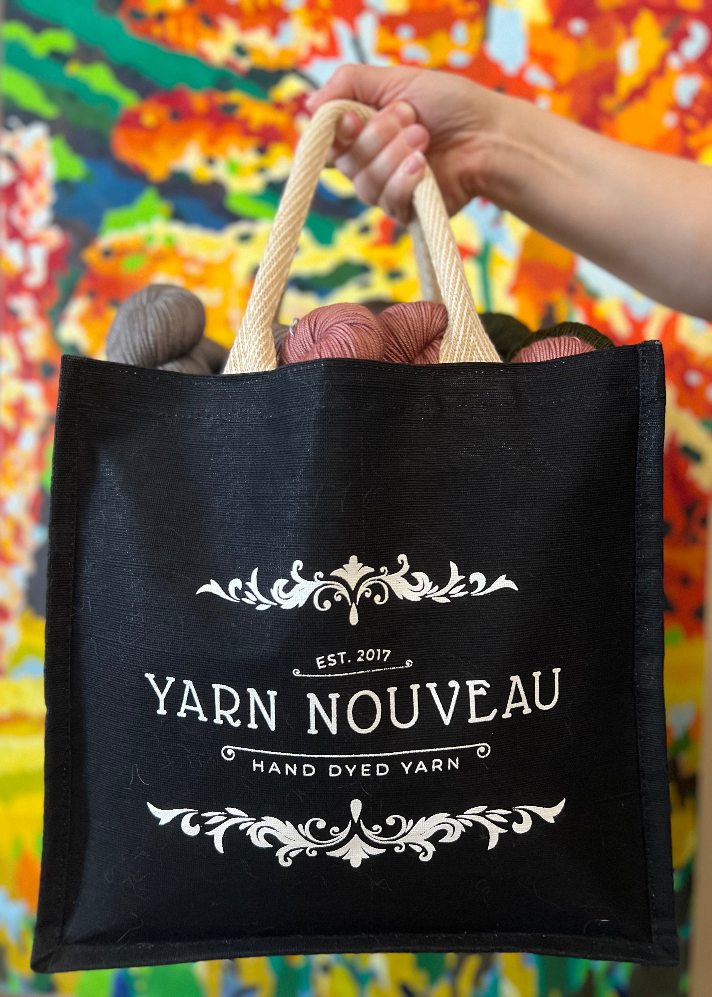 Black tote bag with 'Yarn Nouveau' branding holding yarn against a colorful floral background